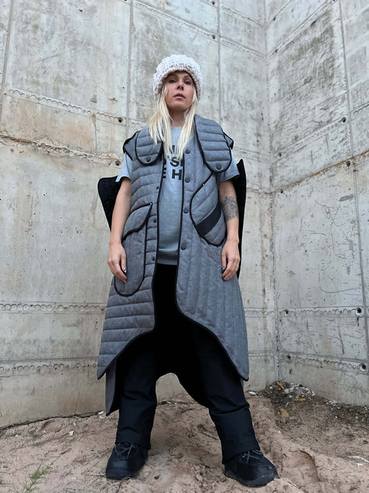 Person wearing a long gray vest over a gray shirt against a concrete wall.