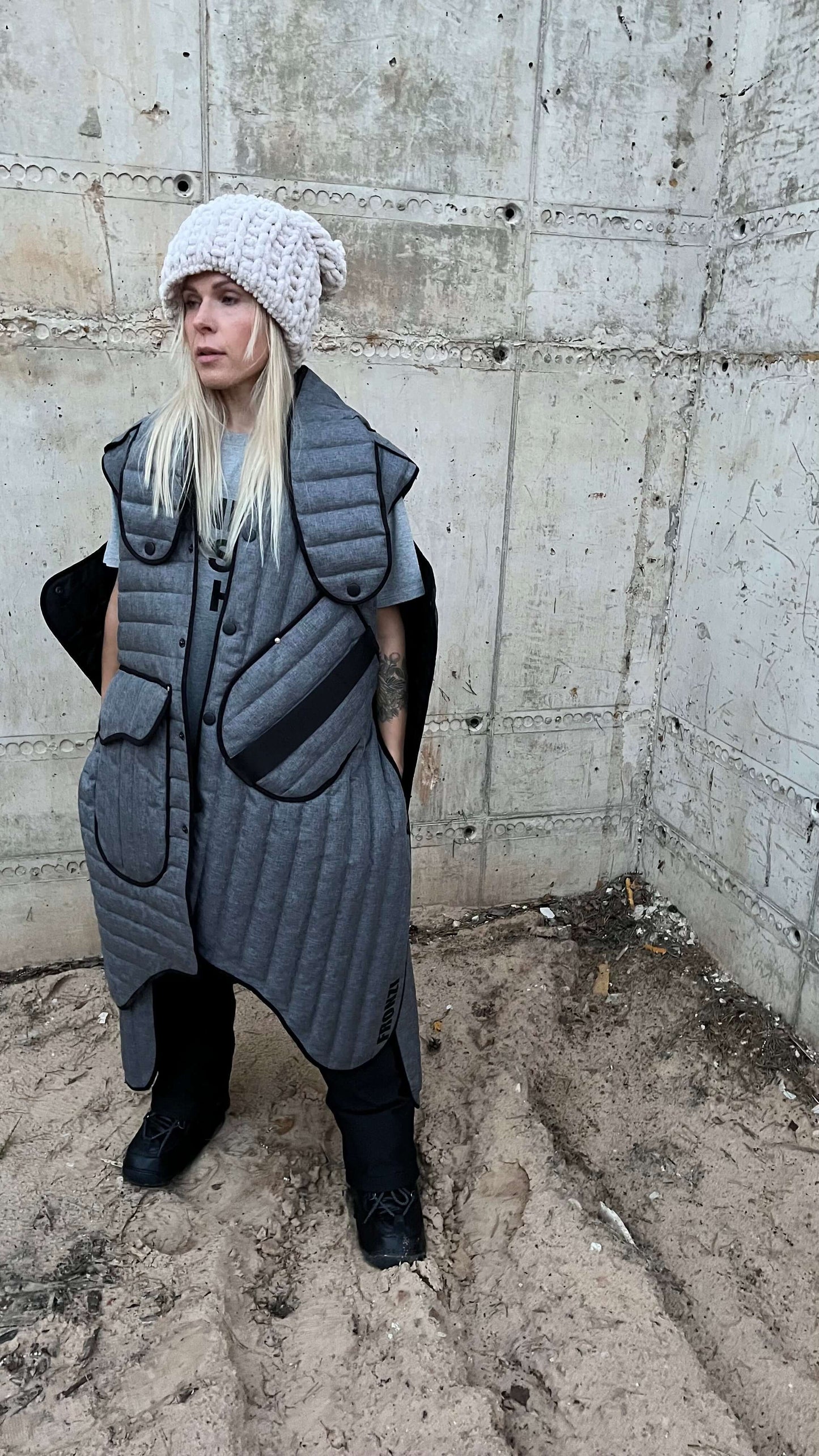 Side view of a blond model wearing a long gray puffer vest and black boots against a concrete wall.