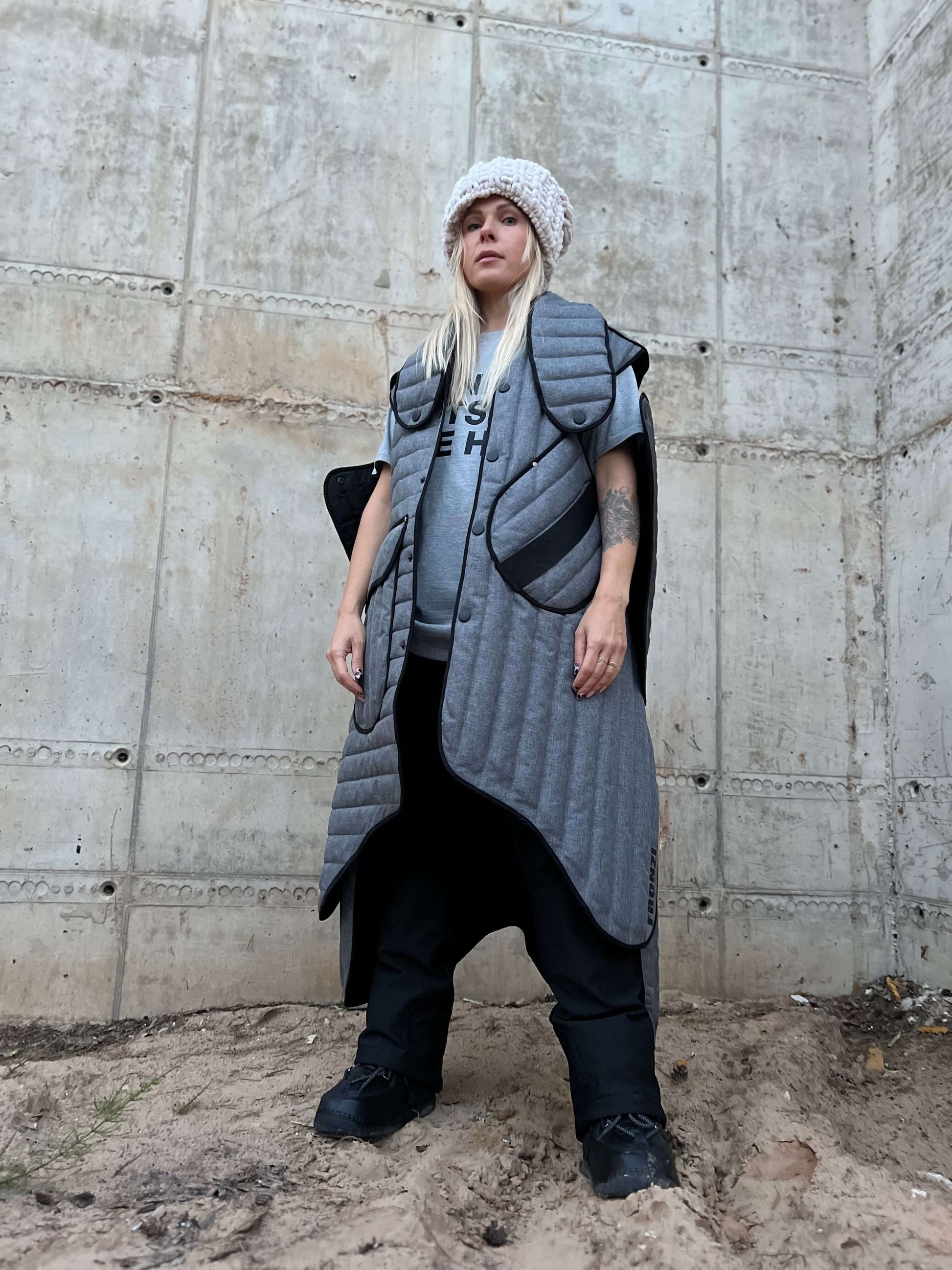 View from lower angle of a blond model wearing a long gray puffer coat against a concrete wall.