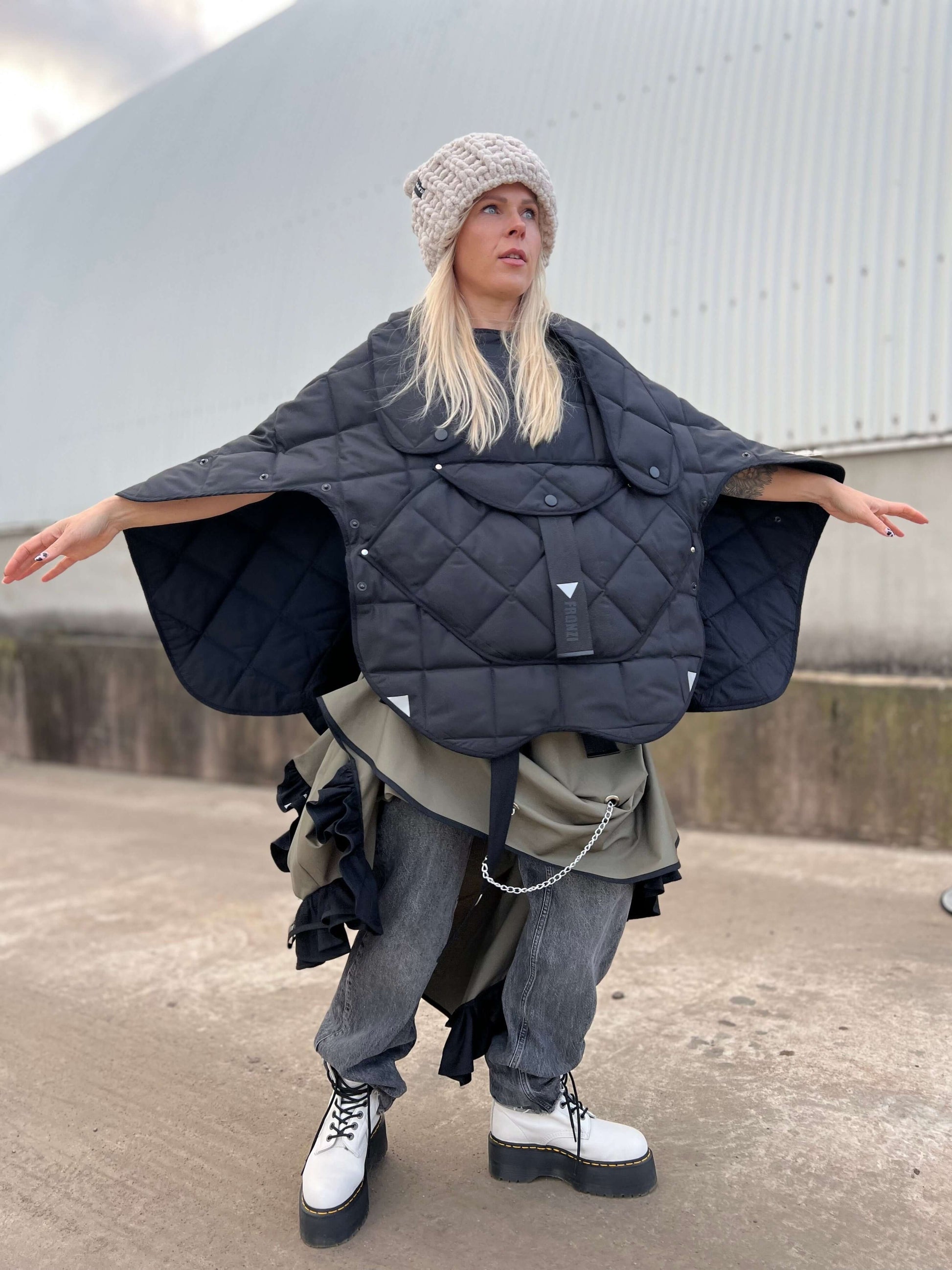 Blond model wearing short quilted hooded jacket with her arms wide open to show that you can wear it as a poncho type of clothing