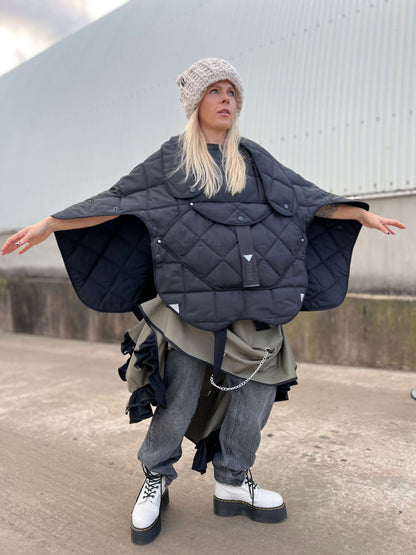 Blond model wearing short quilted hooded jacket with her arms wide open to show that you can wear it as a poncho type of clothing